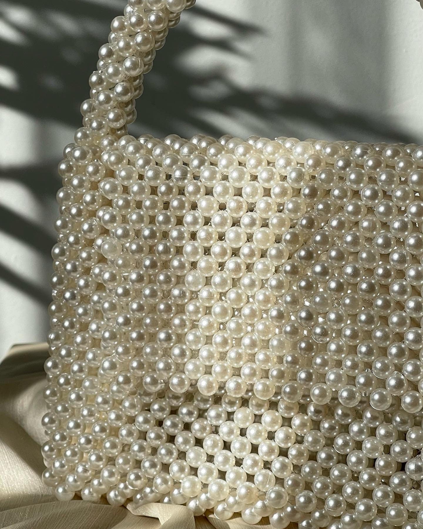 May include: A white pearl beaded handbag with a top handle. The bag is made of small, round pearls that are woven together to create a mesh-like design.