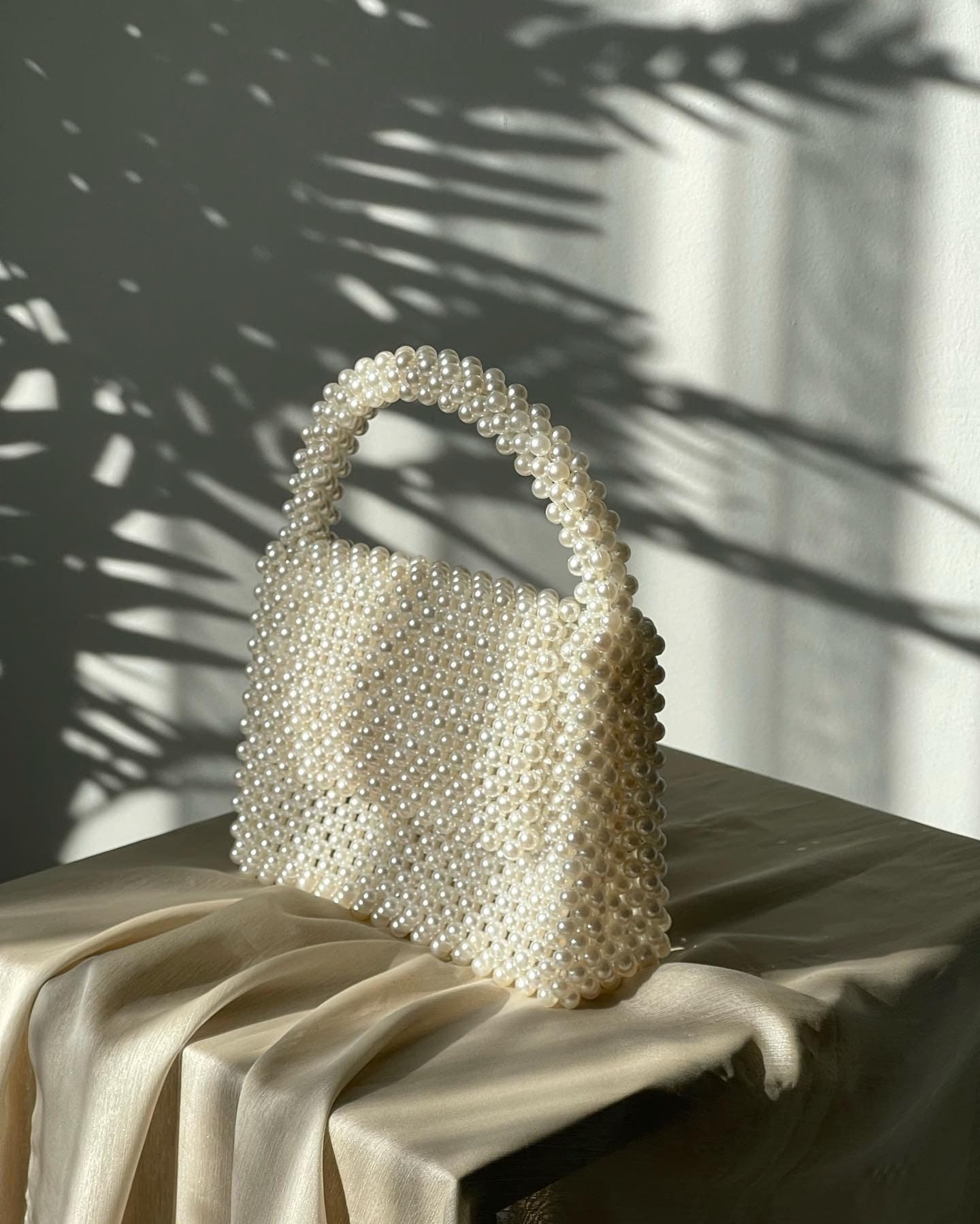 May include: A white beaded handbag with a top handle. The bag is made of small, round pearls and has a rectangular shape.