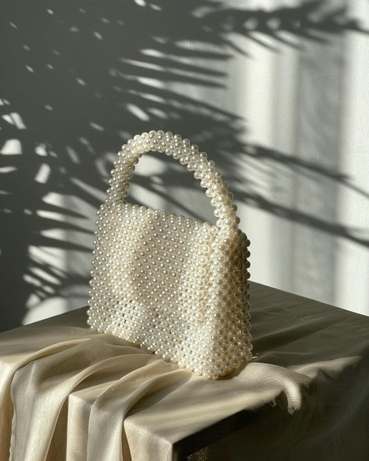 May include: A white beaded handbag with a top handle. The bag is made of small, round pearls and has a rectangular shape.